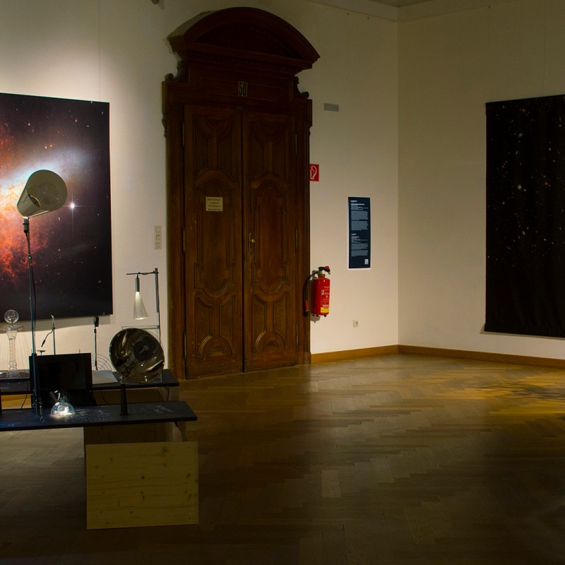 Exhibition room with two large space-themed photographs on the walls and several installations, including a sculpture
                                 on a pedestal. (AI-generated alt-text, generated with GPT-4.1-mini)