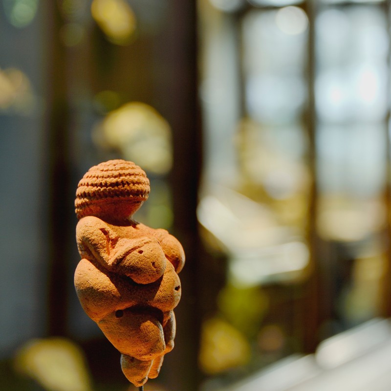 Small prehistoric female figurine with emphasized body features on a stand, blurry museum interior in the background.
                                 (AI-generated alt-text, generated with GPT-4.1-mini)