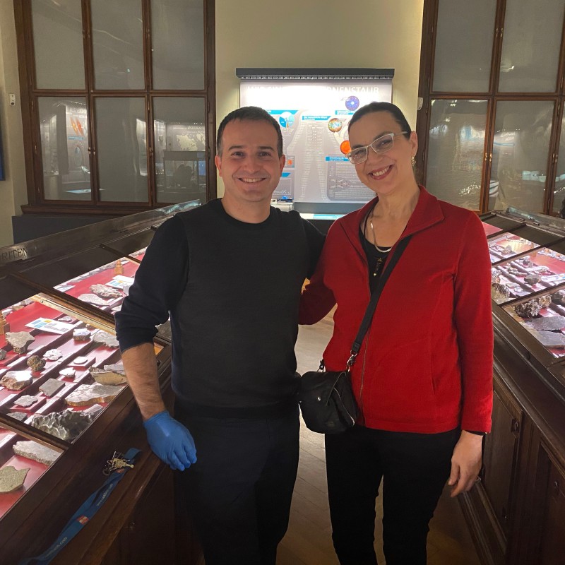 Two people stand inside a museum between display cases with rock samples on red lining. The person on the left wears
                                 black clothing and blue gloves, the person on the right wears a red sweater and glasses. (AI-generated alt-text, generated
                                 with GPT-4.1-mini)