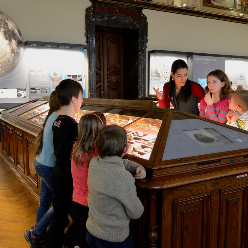 A woman talks to a group of children gathered around a wooden display case in an exhibition room. In the background,
                                 there is a large, round depiction of a celestial body on the wall. (AI-generated alt-text, generated with GPT-4.1-mini)