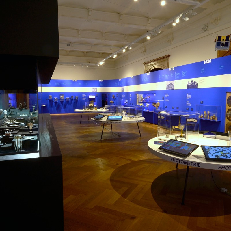 Exhibition room with blue wall, historical instruments in display cases and on white tables, several touchscreens,
                                 and information panels. (AI-generated alt-text, generated with GPT-4.1-mini)