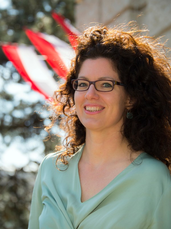 Frau mit lockigen braunen Haaren, Brille
                                 und hellem Oberteil lächelt im Freien, im Hintergrund wehen rot-weiß-rote Fahnen. (KI-generierter Alt-Text, erstellt mit GPT-4.1-mini)