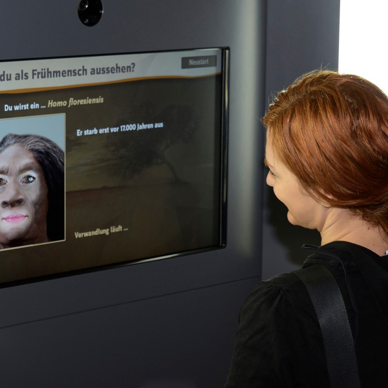 Frau mit rötlichen Haaren betrachtet einen Bildschirm mit einer Rekonstruktion eines Homo floresiensis-Gesichts
                                 und erläuterndem Text. (KI-generierter Alt-Text, erstellt mit GPT-4.1-mini)