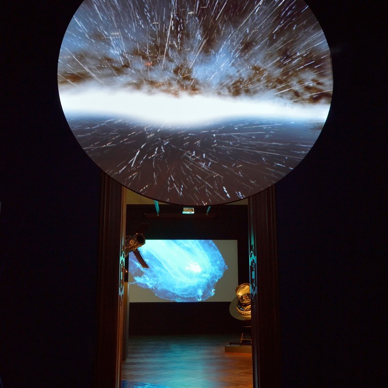 Projektionsinstallation mit kosmischer Nebelwolke und Lichtstrahlen, dahinter Ausstellungsraum mit weiterer Leuchtprojektion.
                                 (KI-generierter Alt-Text, erstellt mit GPT-4.1-mini)