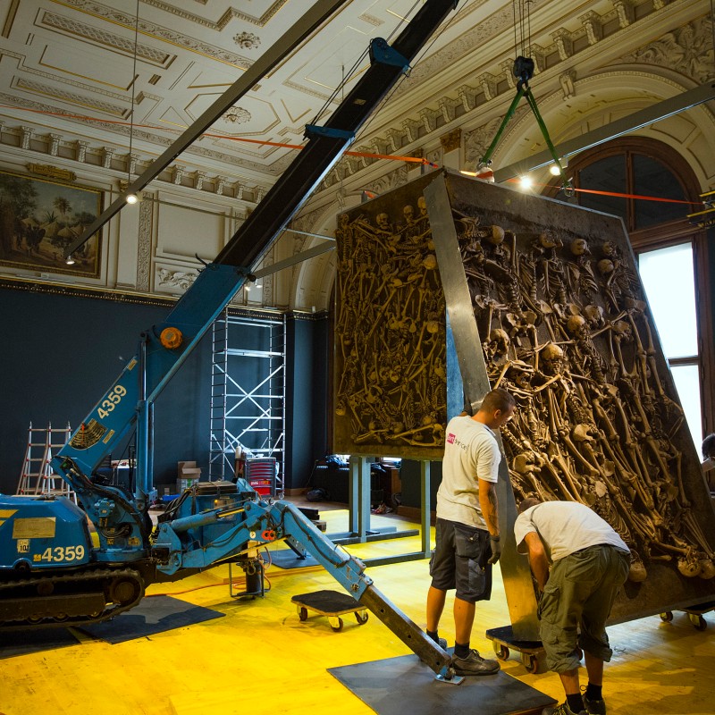 Four men use a crane to move a large rectangular metal structure covered with bones inside a museum room with an
                                 ornate ceiling. (AI-generated alt-text, generated with GPT-4.1-mini)