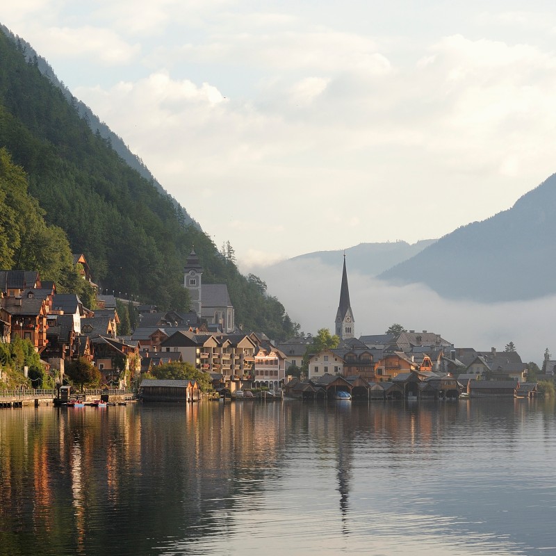 View of a calm lakeshore with picturesque wooden houses, a church tower behind them, and steep forested hillsides
                                 in morning mist. (AI-generated alt-text, generated with GPT-4.1-mini)
