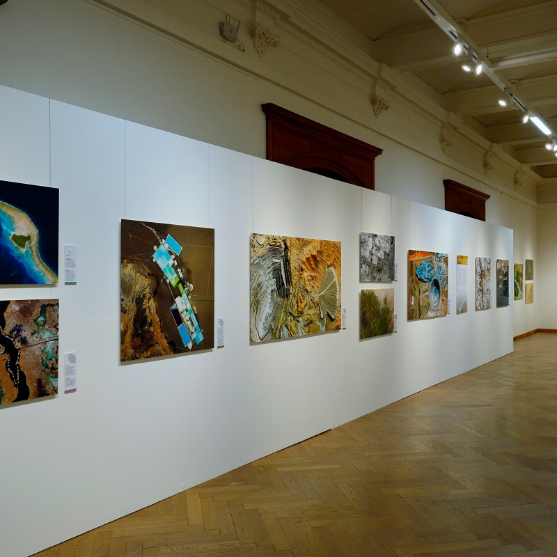 Wide exhibition room with a long white wall displaying multiple color photographs of landscapes and structures.
                                 (AI-generated alt-text, generated with GPT-4.1-mini)
