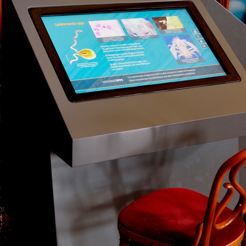 Gray information terminal with large display showing several colored graphic elements and text. A red chair stands
                                 in front. (AI-generated alt-text, generated with GPT-4.1-mini)