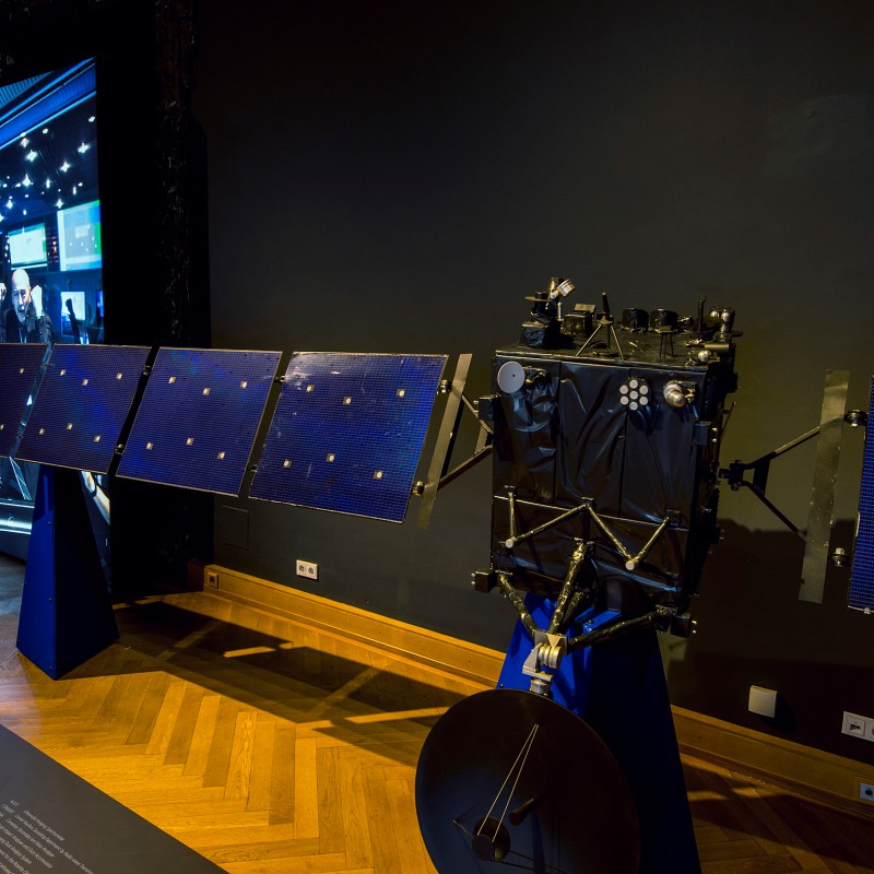 Large satellite model with extended solar panels; group of people on a screen in the background against a dark wall.
                                 (AI-generated alt-text, generated with GPT-4.1-mini)