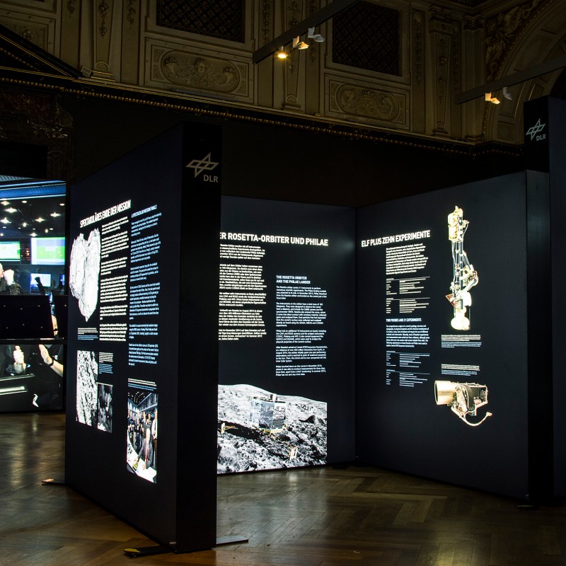 Several black exhibition panels with texts, photos, and graphics about the Rosetta spacecraft in a museum hall.
                                 (AI-generated alt-text, generated with GPT-4.1-mini)