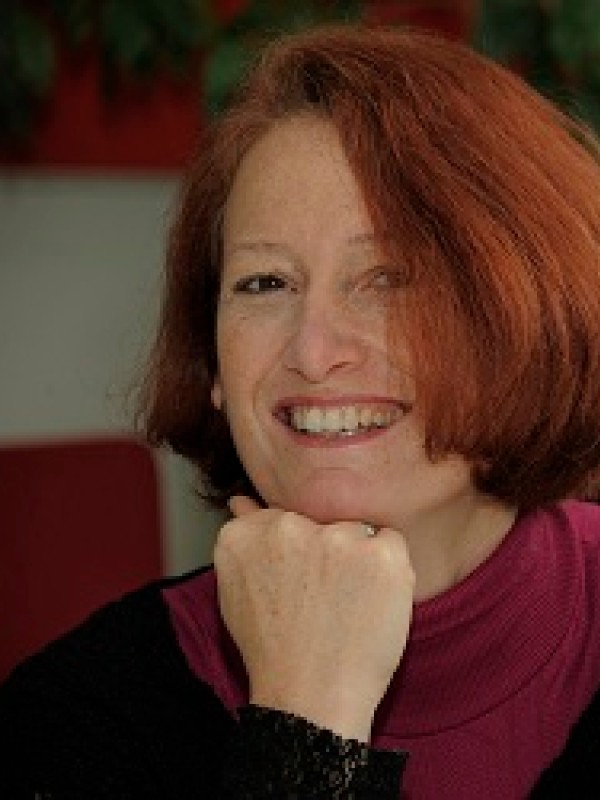 Frau mit kinnlangen roten Haaren lächelt
                                 und hält ein farbig gestaltetes Buch mit der Aufschrift 'NHM TOP 100'. (KI-generierter Alt-Text, erstellt mit GPT-4.1-mini)