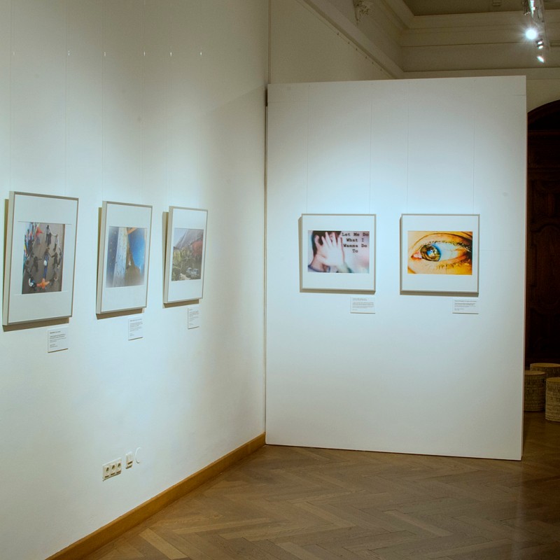 View into an exhibition room with framed photographs on white walls and an area with small stools and round wooden
                                 tables. On the left, a column with clearly legible text: 'Schreibwerkstatt'. (AI-generated alt-text, generated with GPT-4.1-mini)