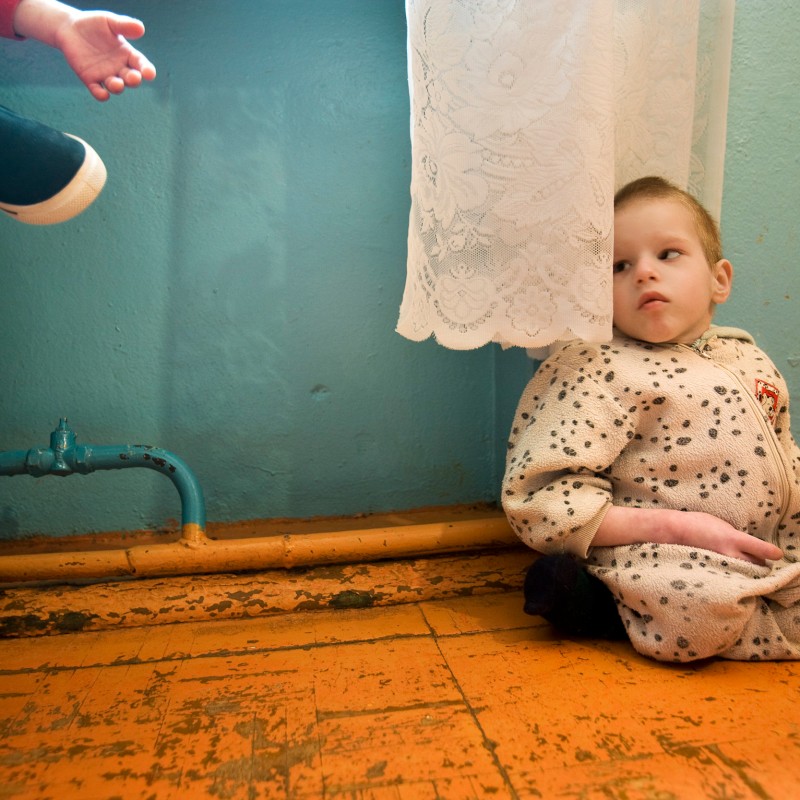 Kleines Kind mit Behinderung sitzt am Boden an einer Wand unter einer weißen Spitzegardine, blickt nach oben zu
                                 einer ausgestreckten Hand. (KI-generierter Alt-Text, erstellt mit GPT-4.1-mini)