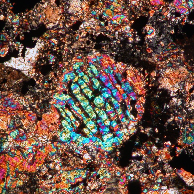 Bunte Mikroaufnahme eines Chondruls aus dem Chelyabinsk-Meteoriten mit vielfarbigen, schillernden Mustern vor dunklem,
                                 körnigem Hintergrund. (KI-generierter Alt-Text, erstellt mit GPT-4.1-mini)