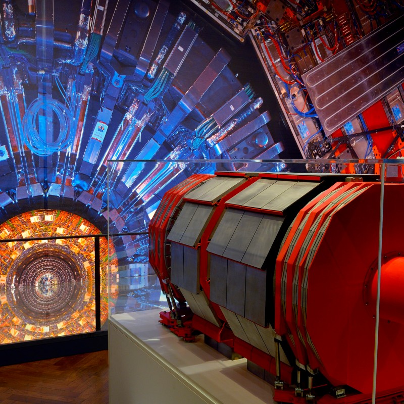 Modell eines roten Teilchendetektors vor einem großen Bild eines komplexen wissenschaftlichen Detektors mit vielen
                                 Kabeln und Leuchtelementen. (KI-generierter Alt-Text, erstellt mit GPT-4.1-mini)