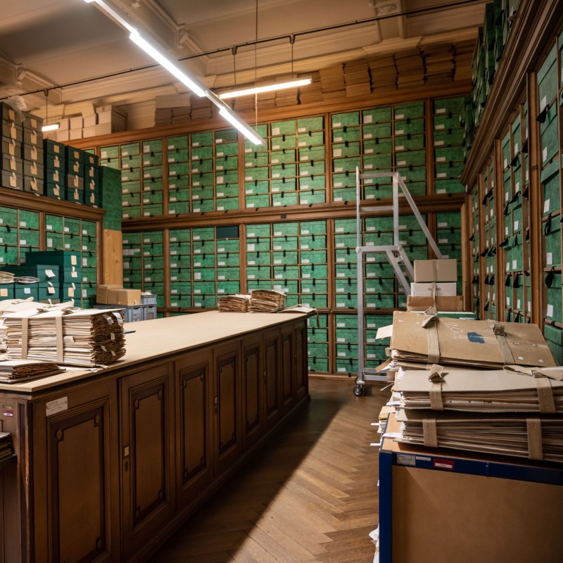 Archivraum mit hohen Regalen voller grüner Archivboxen und einem großen Holztisch mit gestapelten Papierbündeln