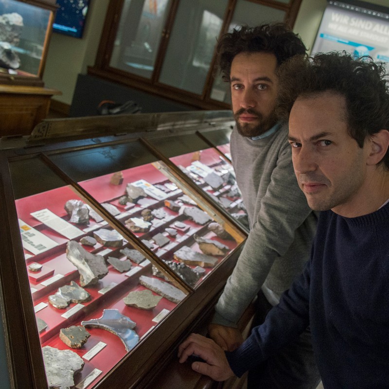 Two men with dark hair looking into a glass display case filled with rock samples in a museum. (AI-generated alt-text,
                                 generated with GPT-4.1-mini)