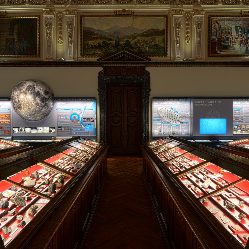 Ornate exhibition room with
                                 multiple display cases containing rock samples and meteorites, illuminated information panels along the walls, large moon
                                 model on the left. (AI-generated alt-text, generated with GPT-4.1-mini)