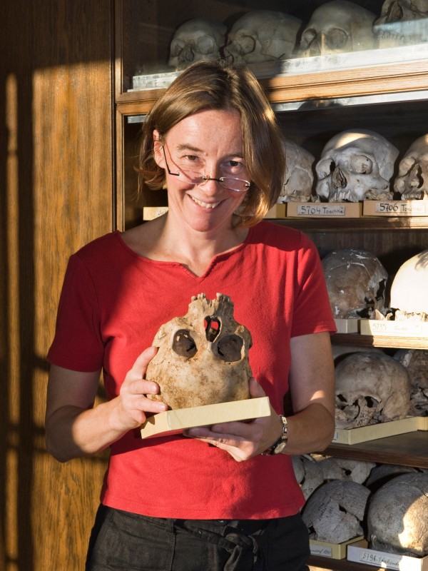 Frau mit Brille und rotem
                                 T-Shirt hält einen menschlichen Schädel, im Hintergrund Regale mit weiteren Schädeln. (KI-generierter Alt-Text, erstellt mit
                                 GPT-4.1-mini)