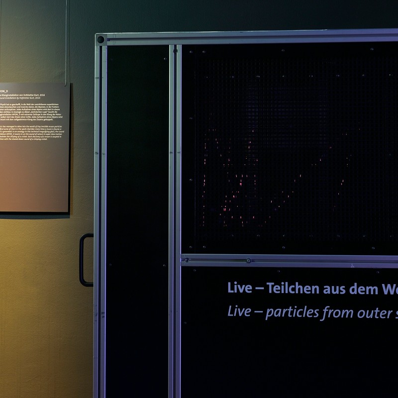 Dunkler Ausstellungsbereich mit einer großen Wandtafel und zwei beleuchteten Informationstafeln mit Text und Grafik.
                                 Auf der Wandtafel steht: 'Live – Teilchen aus dem Weltall'. (KI-generierter Alt-Text, erstellt mit GPT-4.1-mini)