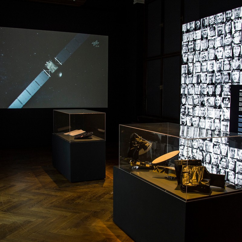 Ausstellungsraum
                                 mit drei Vitrinen, leuchtender Wand mit vielen schwarz-weißen Porträtfotos, großem Bildschirm mit Raumsonde vor Sternenhintergrund.
                                 (KI-generierter Alt-Text, erstellt mit GPT-4.1-mini)