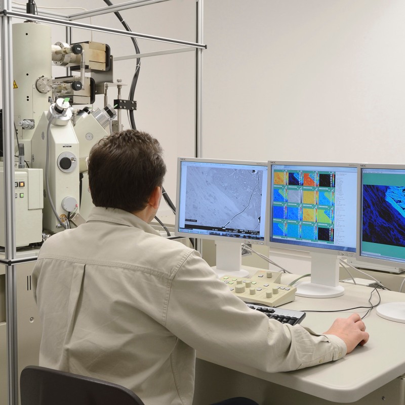 Man with short dark hair sits at a control panel in front of multiple monitors and an electron microscope, screens display
                                 scientific analysis data and graphics. (AI-generated alt-text, generated with GPT-4.1-mini)