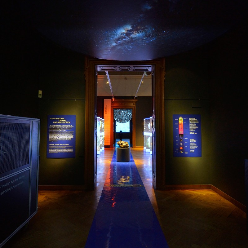 Dunkler
                                 Ausstellungsraum mit beleuchteten Texttafeln an den Wänden, einem Exponat auf einem Podest im Hintergrund und einer blauen
                                 Laufbahn auf dem Boden. (KI-generierter Alt-Text, erstellt mit GPT-4.1-mini)