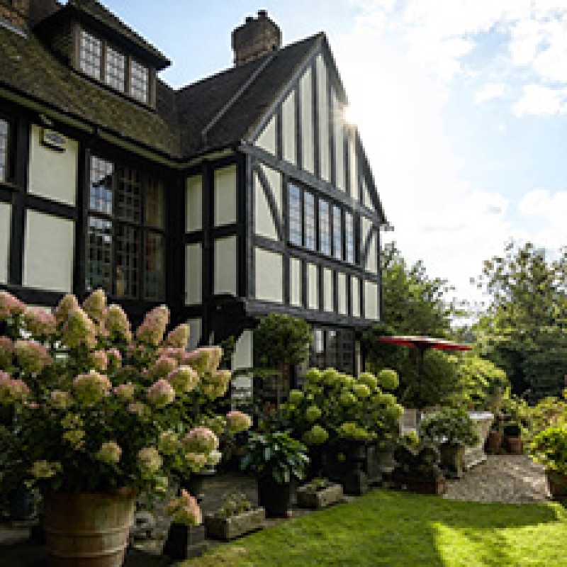 Großes Fachwerkhaus mit markanten dunklen Holzbalken, breiten Fenstern und einem weitläufigen Garten mit üppigen
                                 Sträuchern und Blumen im Vordergrund. (KI-generierter Alt-Text, erstellt mit GPT-4.1-mini)