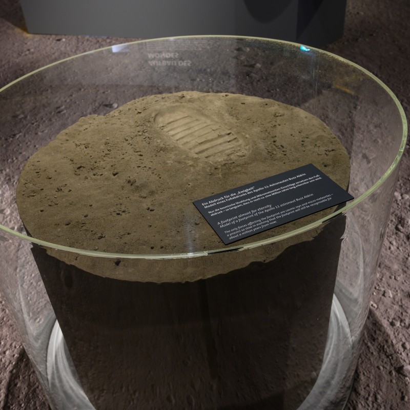 Cylindrical exhibit made of sand-colored material with a single shoe print, displayed under a round glass cover.
                                 A dark info plaque with white text sits on top side. (AI-generated alt-text, generated with GPT-4.1-mini)