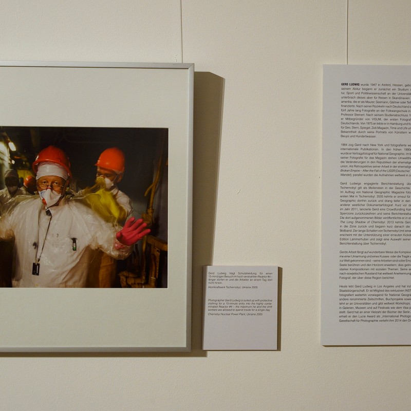 Photograph of people in protective suits and helmets with pink gloves, next to a wall text with several paragraphs.
                                 (AI-generated alt-text, generated with GPT-4.1-mini)