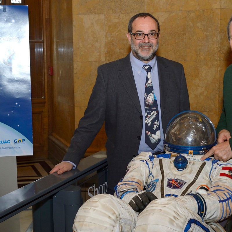 Two men stand beside an astronaut suit, one touching the helmet, next to a poster with an astronaut figure and text
                                 about Mission X. (AI-generated alt-text, generated with GPT-4.1-mini)