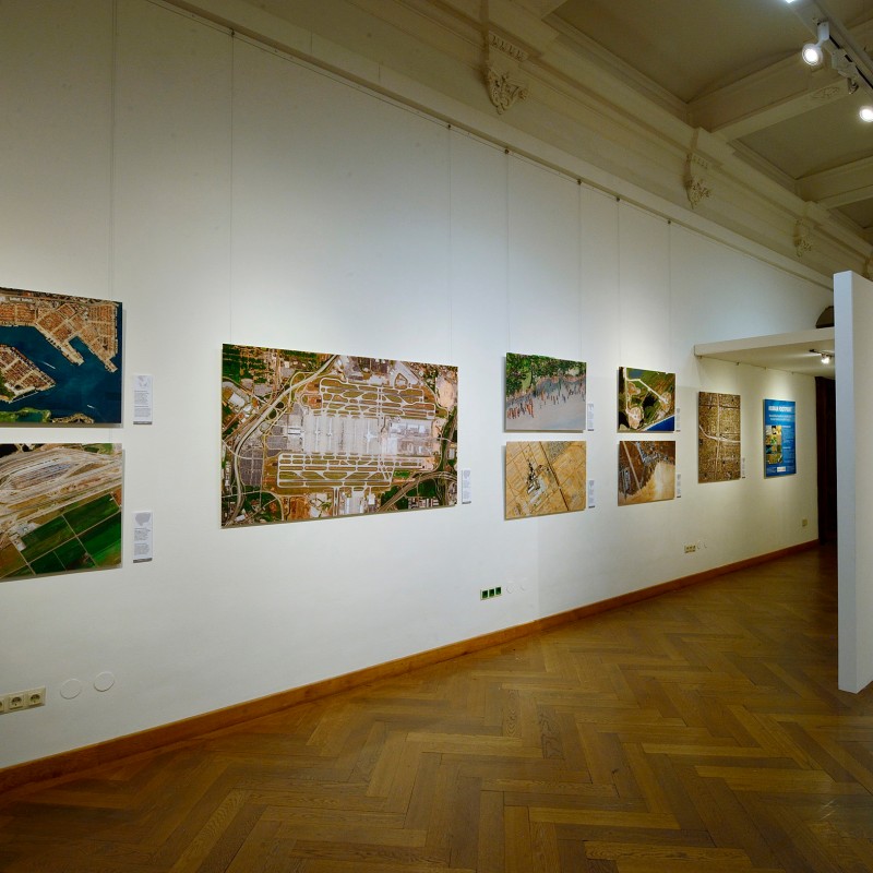 Exhibition room with multiple large aerial photographs on white walls and parquet floor. (AI-generated alt-text,
                                 generated with GPT-4.1-mini)