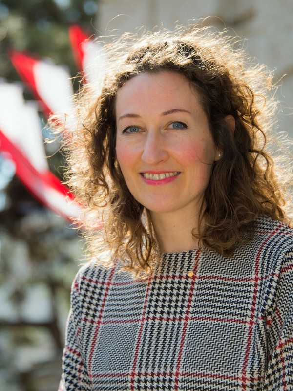 Frau mit lockigen,
                                 braunen Haaren und gemustertem Oberteil steht lächelnd draußen vor unscharfem Hintergrund mit roten und weißen Farben. (KI-generierter
                                 Alt-Text, erstellt mit GPT-4.1-mini)