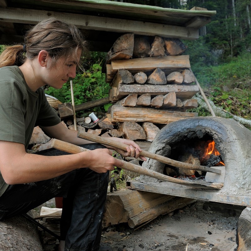 Person with long hair uses wooden sticks to push something into a small clay oven with fire. Stacked firewood and
                                 forest in the background. (AI-generated alt-text, generated with GPT-4.1-mini)