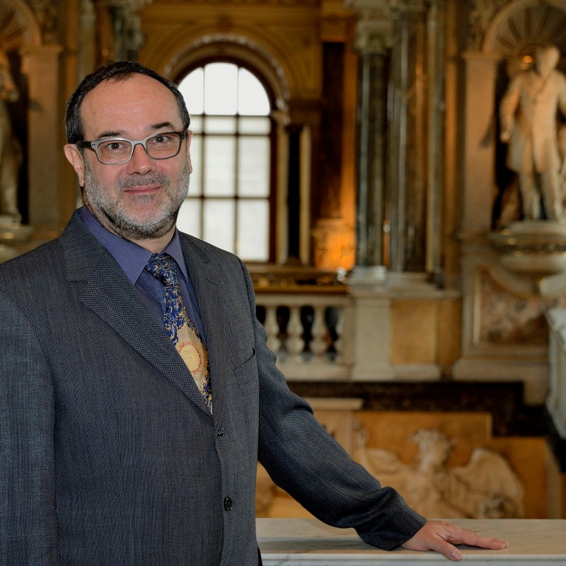 Mann mit Bart, Brille und Anzug steht lächelnd in einer prunkvollen Halle vor marmorner Treppe und Statuen. (KI-generierter
                                 Alt-Text, erstellt mit GPT-4.1-mini)