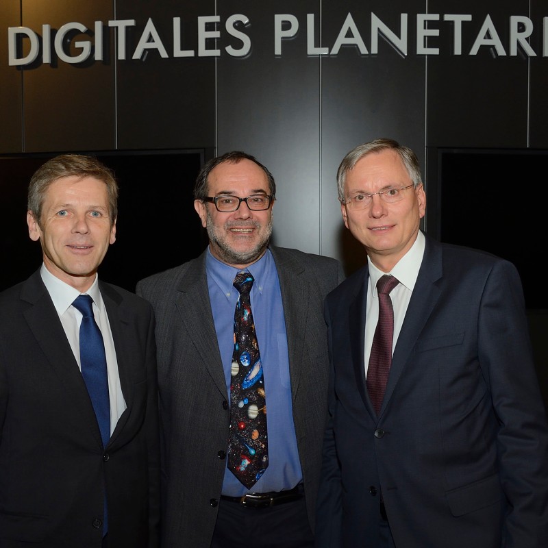 Drei Männer in Anzügen stehen nebeneinander und lächeln vor einer Wand mit der Aufschrift 'DIGITALES PLANETARIUM'.
                                 (KI-generierter Alt-Text, erstellt mit GPT-4.1-mini)
