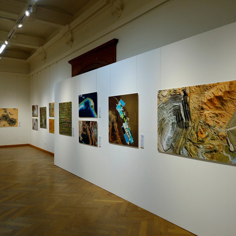 View into an exhibition hall with large-format aerial and satellite images of human impacts on landscapes, displayed
                                 on white walls. (AI-generated alt-text, generated with GPT-4.1-mini)