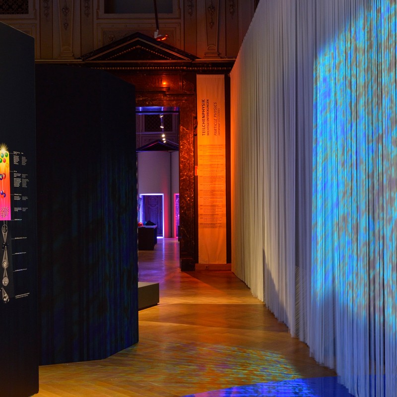 Blick
                                 in einen abgedunkelten Ausstellungsraum mit Lichtprojektionen auf einen weißen Vorhang und Infotafeln an einer schwarzen Wand.
                                 (KI-generierter Alt-Text, erstellt mit GPT-4.1-mini)