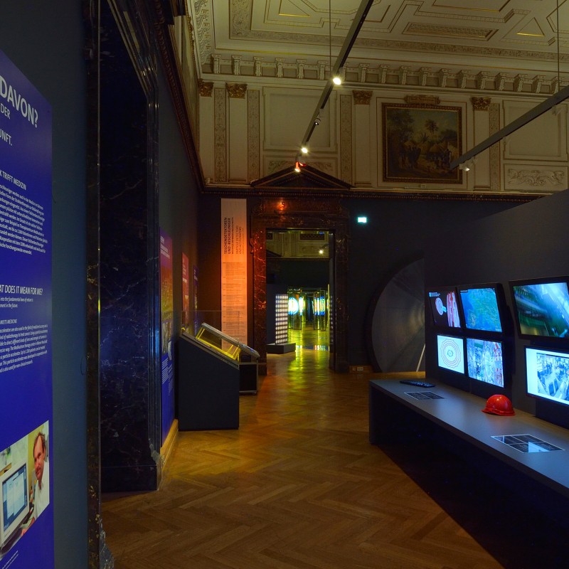 Museumsausstellungsraum
                                 mit Holzfußboden, hohen verzierten Decken, beleuchteten Wandtafeln links sowie mehreren Monitoren auf einer langen, dunklen
                                 Konsole rechts. Zwei Schutzhelme stehen nebeneinander auf der Konsole. (KI-generierter Alt-Text, erstellt mit GPT-4.1-mini)