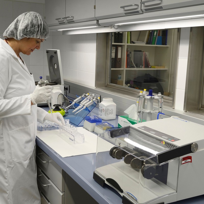 Researcher in a white lab coat and hairnet stands at a laboratory bench with equipment, holding a pipette. (AI-generated
                                 alt-text, generated with GPT-4.1-mini)