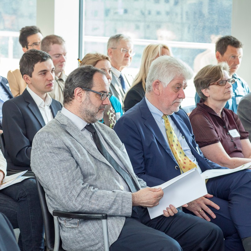 Multiple people sit close together in a bright room, listening attentively; some hold papers in their hands. (AI-generated
                                 alt-text, generated with GPT-4.1-mini)