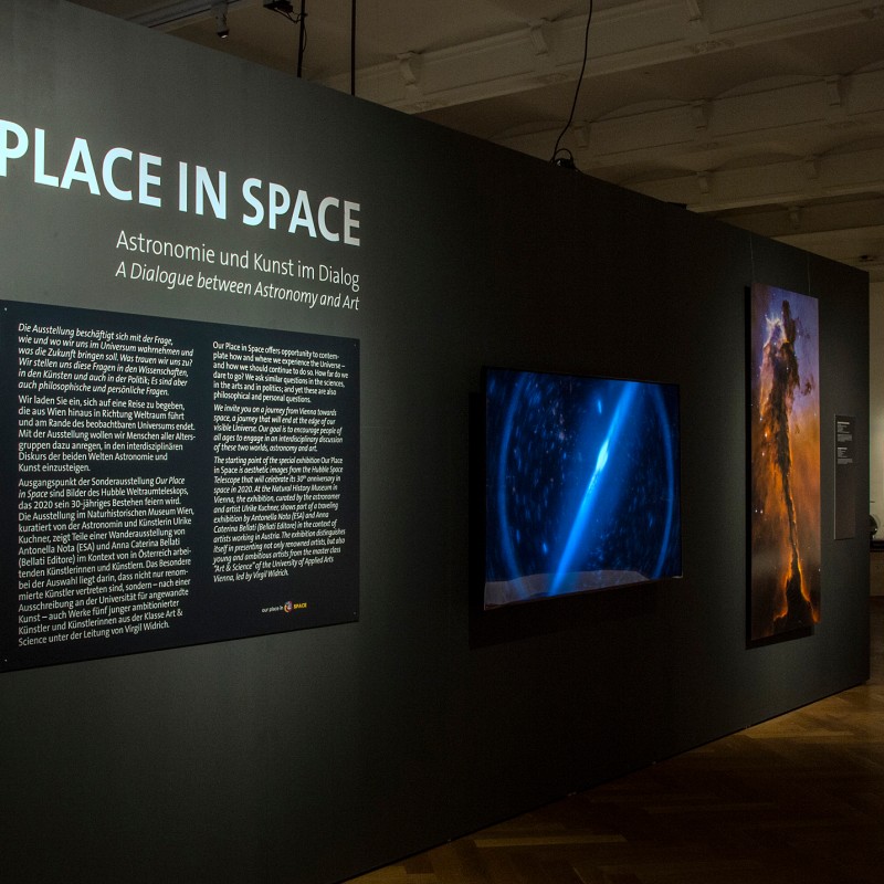 Ausstellungstafel mit dem Titel
                                 'OUR PLACE IN SPACE' und Erklärungstext auf Deutsch und Englisch an einer Wand, daneben drei großformatige Bilder mit Weltraummotiven
                                 in einem Museum (KI-generierter Alt-Text, erstellt mit GPT-4.1-mini)