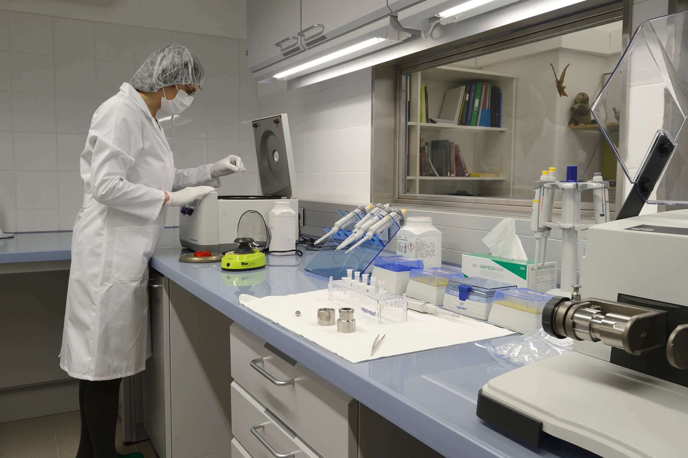 Cleanroom: Person im weißen Laborkittel und Haarnetz arbeitet an einem Labortisch im Reinraum; Laborgeräte, Reagenzgläser und Zentrifuge sichtbar. (KI-generierter Alt-Text, erstellt mit GPT-4.1-mini)