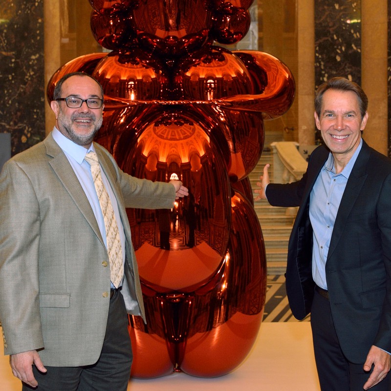 Zwei Männer im Anzug stehen lächelnd vor einer großen, glänzenden, orangefarbenen Skulptur in einem historischen
                                 Saal mit Marmorsäulen und gemustertem Boden. (KI-generierter Alt-Text, erstellt mit GPT-4.1-mini)