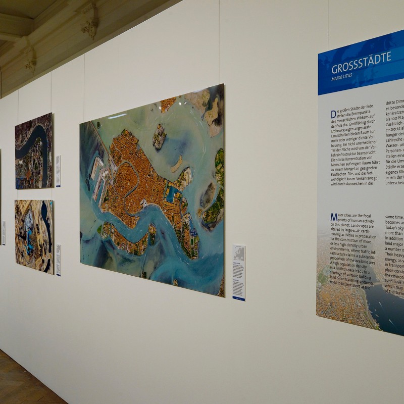 Exhibition wall displaying multiple large-format aerial photographs of cities next to an information poster with
                                 blue border. (AI-generated alt-text, generated with GPT-4.1-mini)