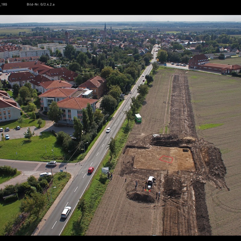 Aerial view of an archaeological excavation site beside a country road at the city outskirts, with multiple cars
                                 on the road and buildings in the background. (AI-generated alt-text, generated with GPT-4.1-mini)