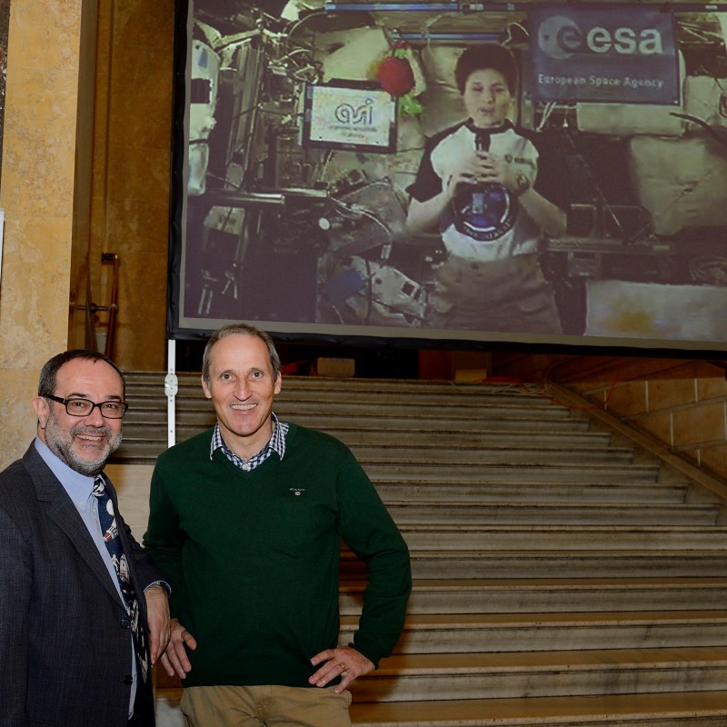 Two men stand on stairs, with a large video projection in the background showing a person in a spacesuit and a visible
                                 ESA logo. (AI-generated alt-text, generated with GPT-4.1-mini)