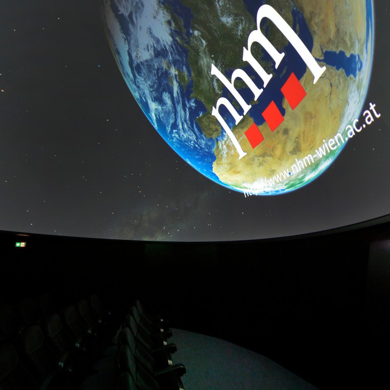 Dimly lit domed room with empty rows of seats and a projection
                                 of a large globe displaying 'nhm' and a website address. (AI-generated alt-text, generated with GPT-4.1-mini)