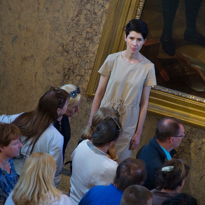 Frau mit kurzem dunklem Haar und beigefarbenem Kleid spricht zu einer Gruppe von Personen vor einem großen, goldgerahmten
                                 Gemälde an einer Wand. (KI-generierter Alt-Text, erstellt mit GPT-4.1-mini)