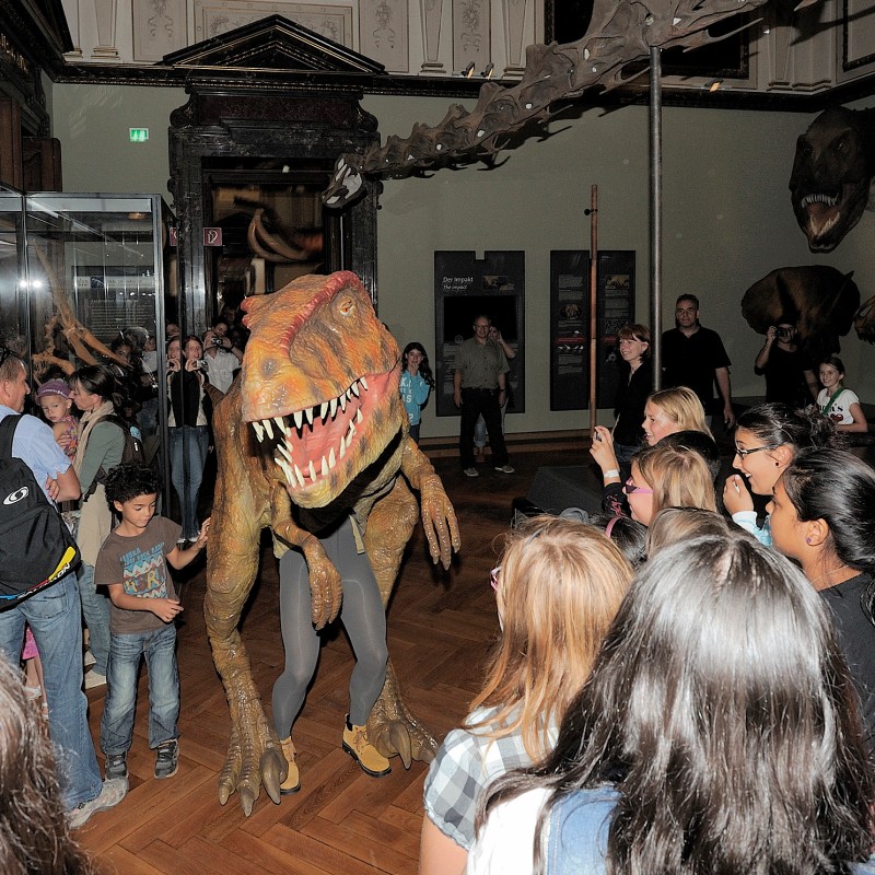 Children and adults gather around a life-sized dinosaur model in a museum hall with skeletal displays on the walls.
                                 (AI-generated alt-text, generated with GPT-4.1-mini)
